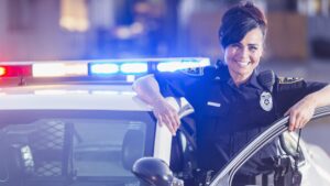 A female police officer getting out of her patrol car. She is smiling, looking at the camera, leaning one arm on the open door of the vehicle. The lights are on.