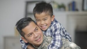 A military dad and his son are hugging in their living room. The son is smiling happily at the camera.