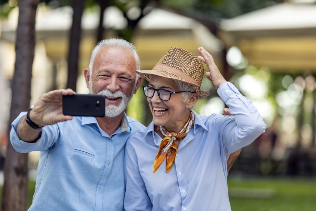 Senior Casually Dressed Couple with Grey Hair is Having Nice Time on Warm Sunny Day in Public Park. They are Sitting on the Wooden Bench and Taking Selfie Photos with Mobile Phone. | Un couple senior habillé de façon décontractée avec des cheveux gris passe du bon temps lors d'une chaude journée ensoleillée dans un parc public. Ils sont assis sur le banc en bois et prennent des photos de selfie avec leur téléphone portable.