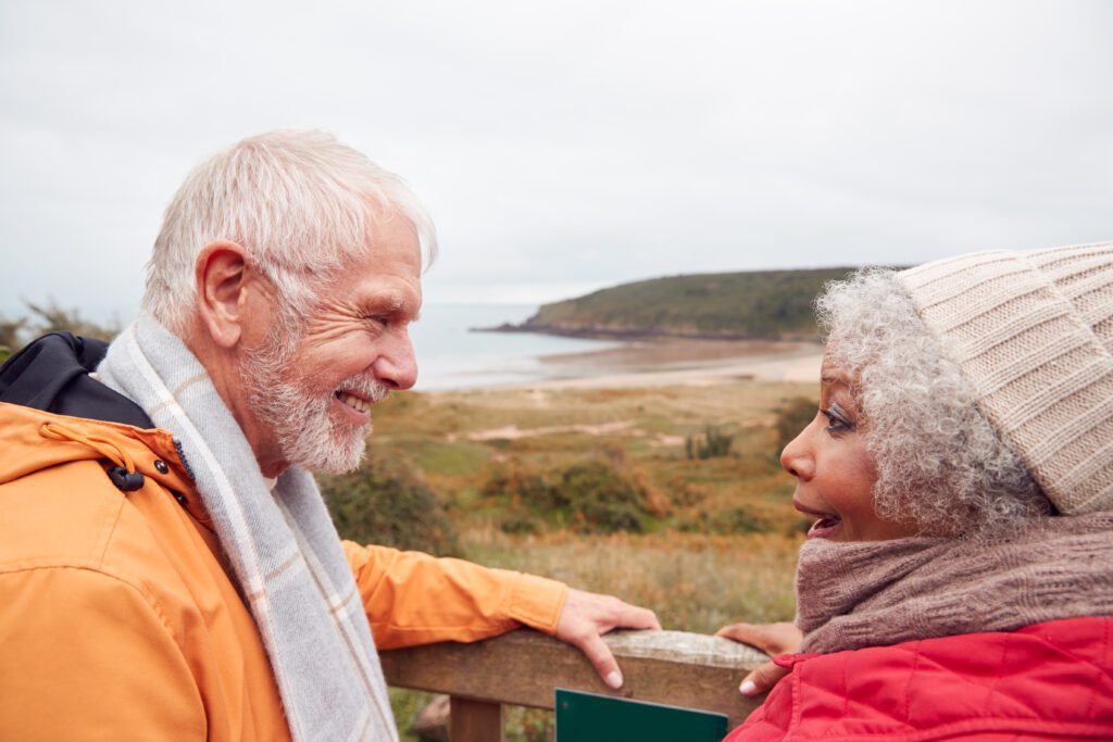 Rear View Of Active Senior Couple Looking Out Over Gate As They Walk Along Coastal Path In Fall | Vue arrière d'un couple senior actif regardant par-dessus la porte alors qu'ils marchent le long du sentier côtier à l'automne