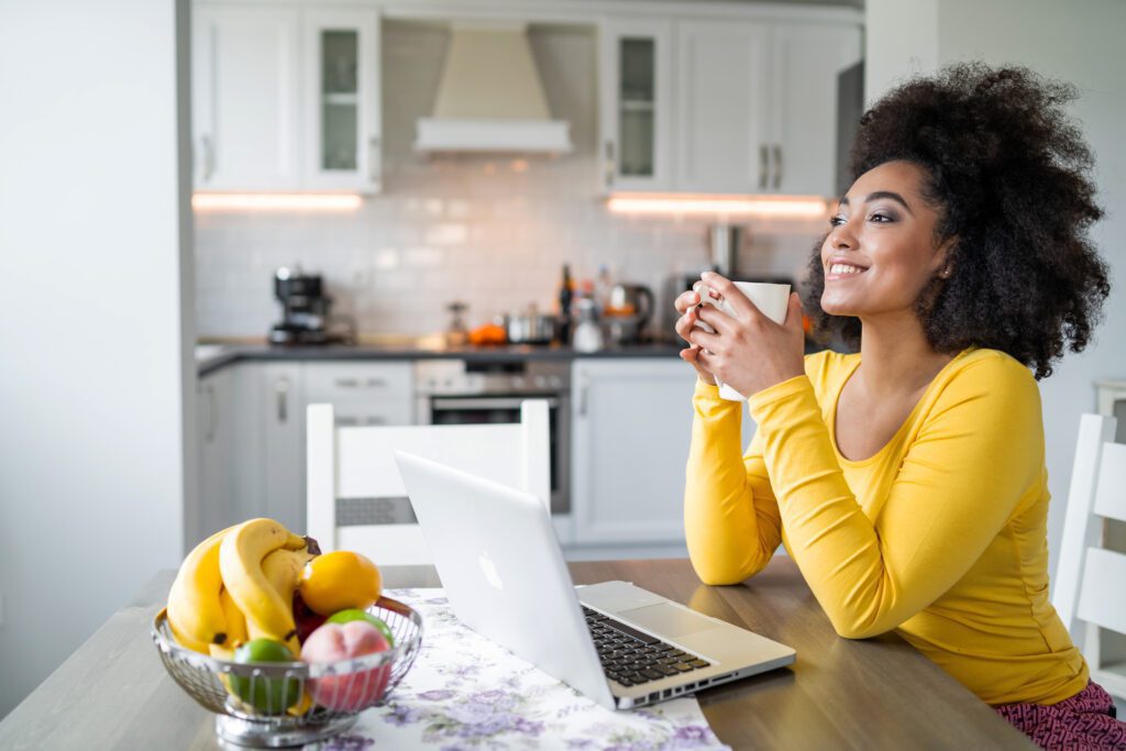Beautiful afro young woman with bright yellow shirt sitting at home, smiling in front of her laptop, having online conversation
