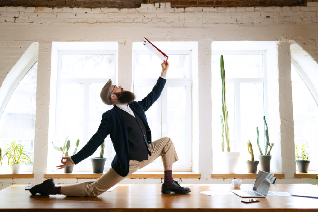 Unusual business process. Young bearded man, office clerk having fun, doing yoga on wooden table in modern office at work time with gadgets. Concept of business, healthy lifestyle, sport, hobby