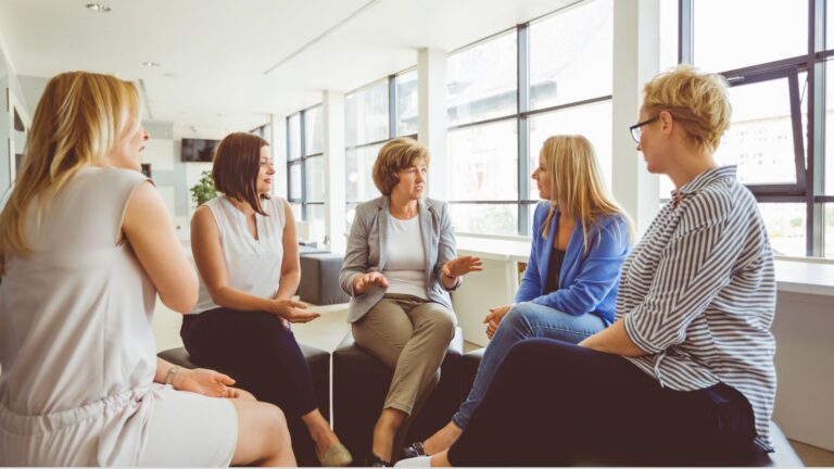 A woman stands in workplace office settings, confidently presenting to an engaged audience amidst a backdrop of books.