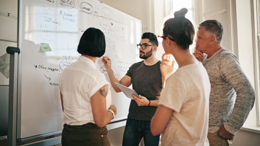 A diverse group of individuals gathered around a whiteboard, engaged in discussion and collaboration.