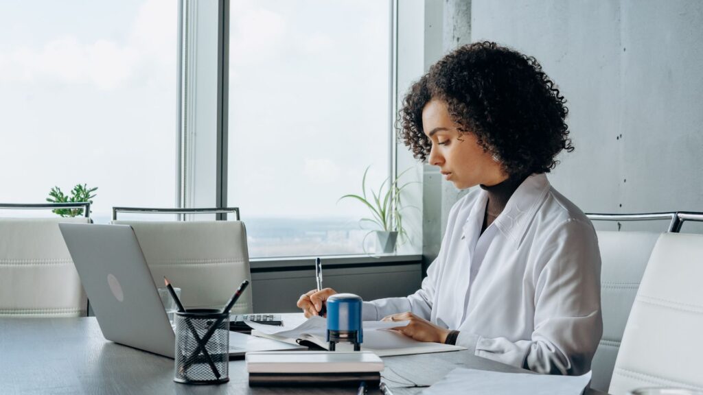 A woman in a white lab coat sits at a desk, working on a laptop in a professional setting