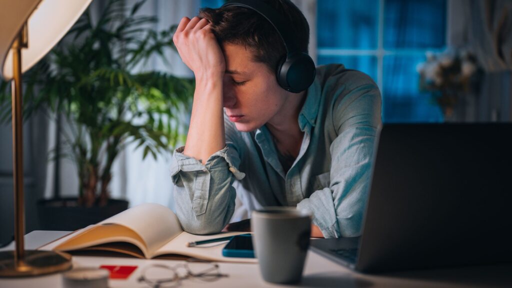 A man wearing headphones sits at a desk with a laptop and an open book in front of him.