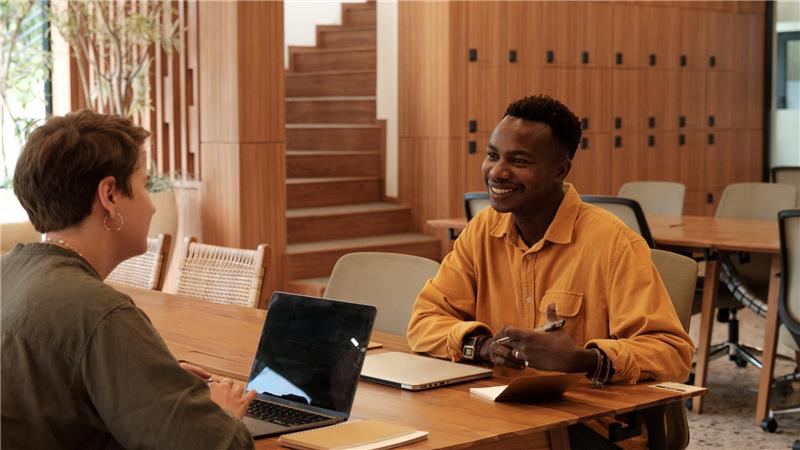 Two men seated at a table, each using a laptop, engaged in conversation or collaboration.
