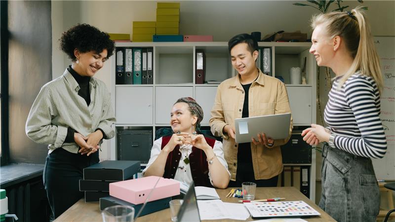 Three individuals gather around a table, engaged in discussion while looking at a laptop screen.