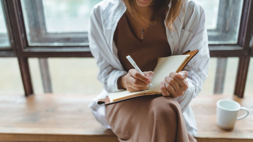 A woman sits on a window sill, writing in a notebook with a pen, surrounded by natural light.