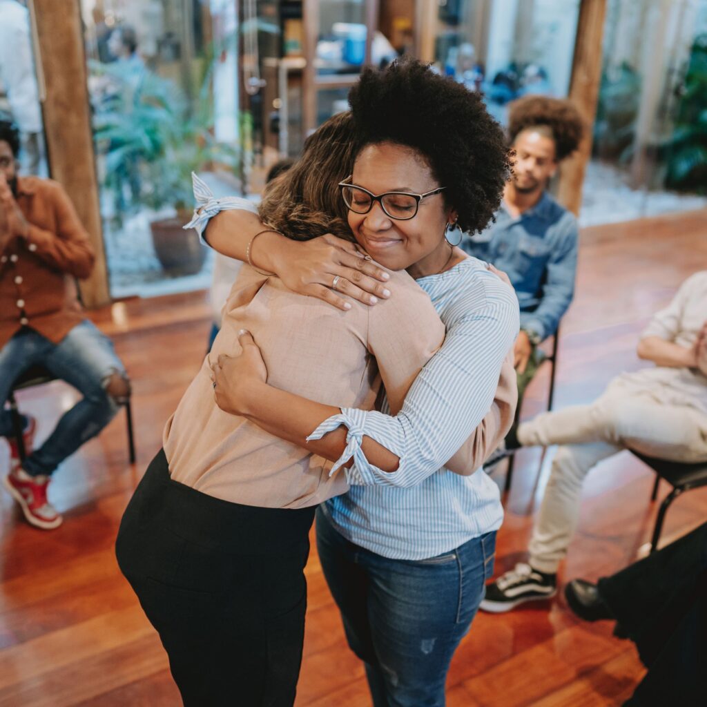 Two women embrace in during mental health training, surrounded by seated colleagues engaged in peer support discussion.
