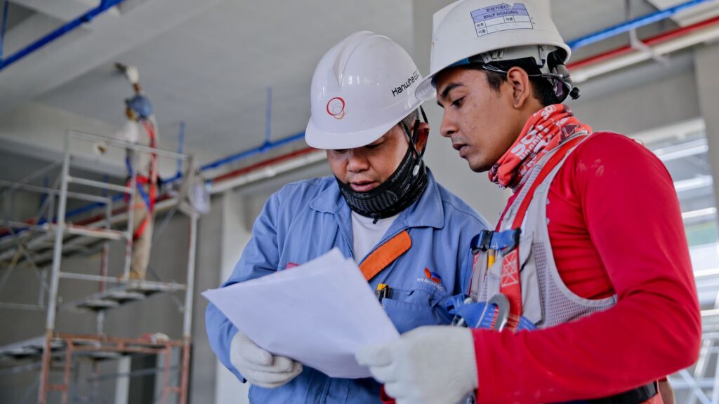 Two workers in safety helmets reviewing plans at a worksite, illustrating occupational health and safety