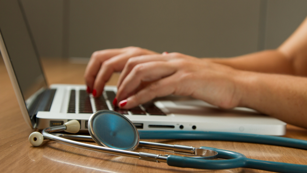 Healthcare professional typing on a laptop next to a stethoscope
