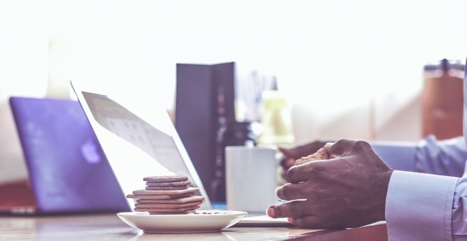 Hands holding coffee with biscuits on desk beside laptop during work break.